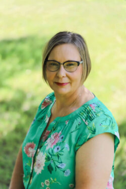 Headshot of Kathy Magnusson, a woman with a chin-length blond bob wearing black framed glasses and a green floral blouse