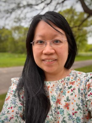 Headshot of Maria Moua, a woman with long black hair wearing unframed glasses and a long-sleeved floral print blouse.