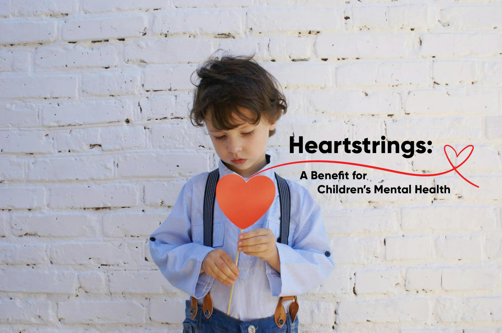 A young boy in suspenders holds a heart-shaped paper against a white brick wall, conveying innocence and emotion.